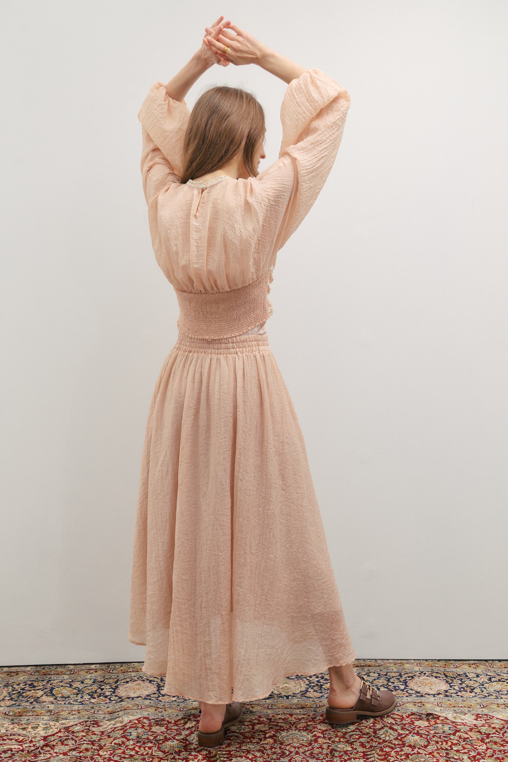 Woman in a long dusty pink skirt and matching top standing with arms raised against a plain wall.