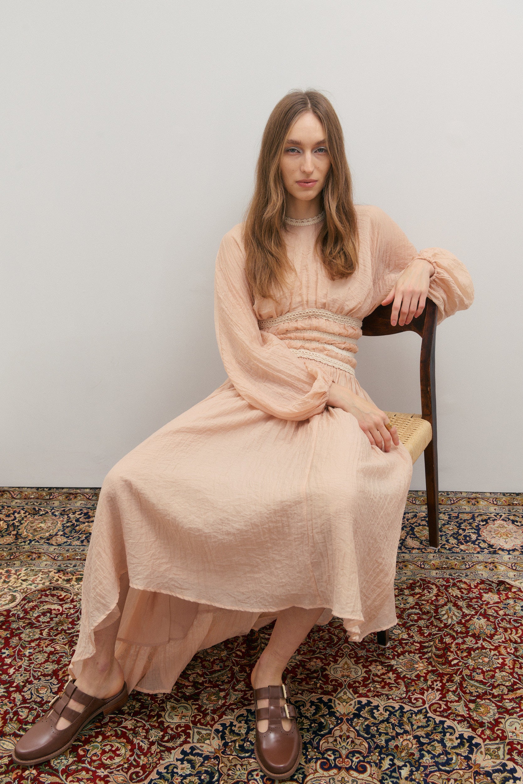 Woman in a long, light pink skirt and matching top sitting on a chair against a plain wall.