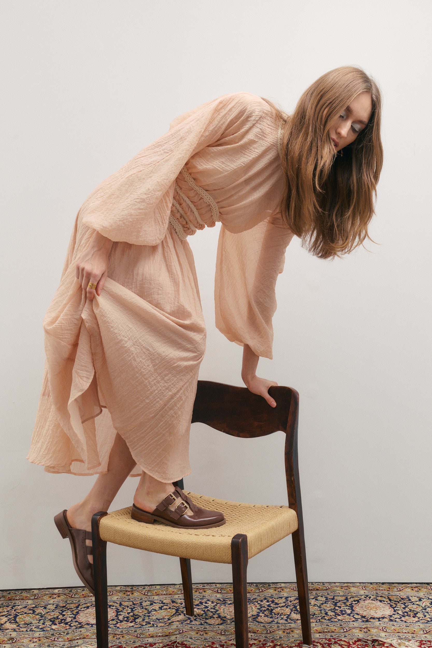 Woman in a light pink blush colour skirt and matching top climbing onto a chair against a plain wall.