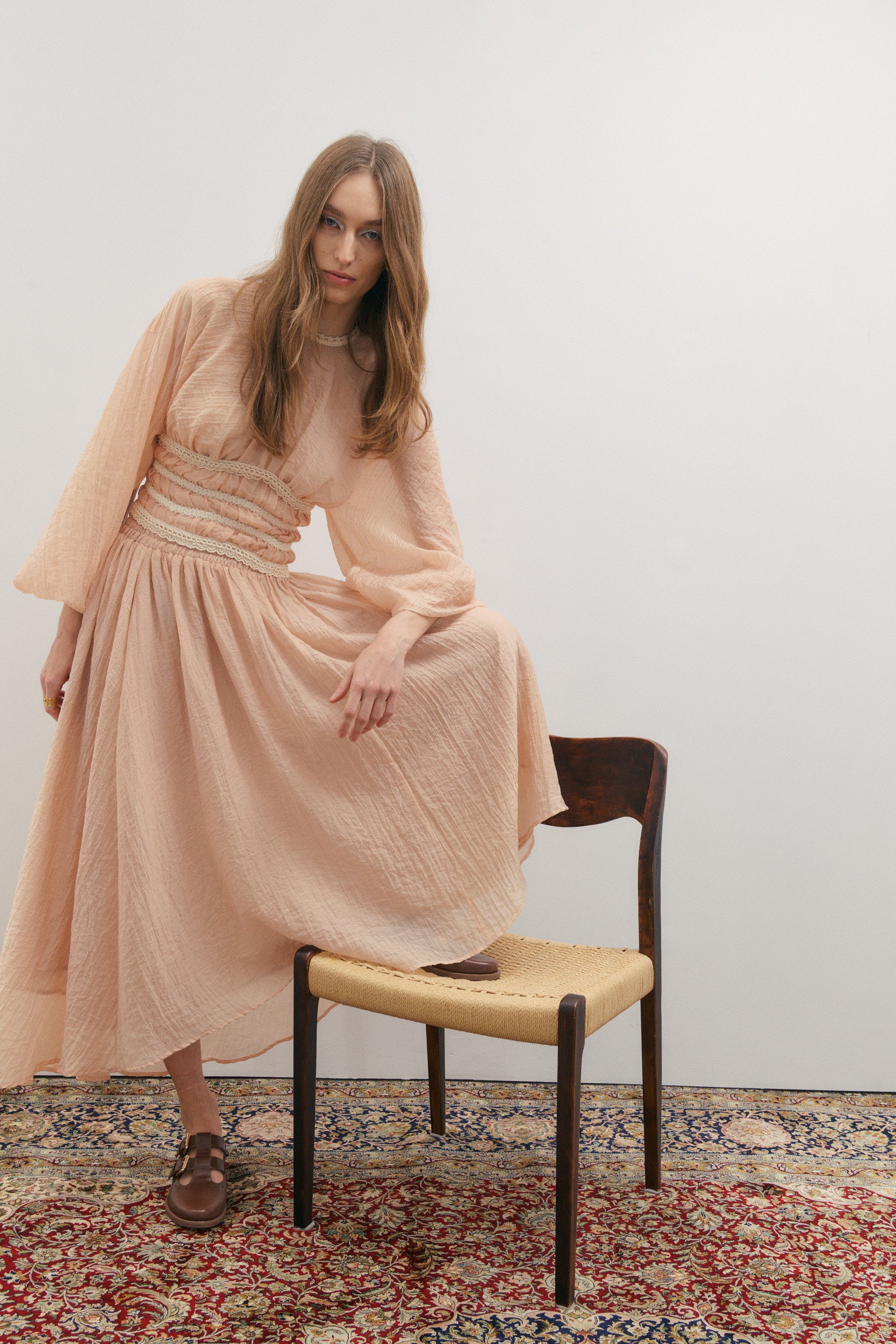 Woman in a long dusty pink skirt and matching top sitting on a wooden chair against a white wall.
