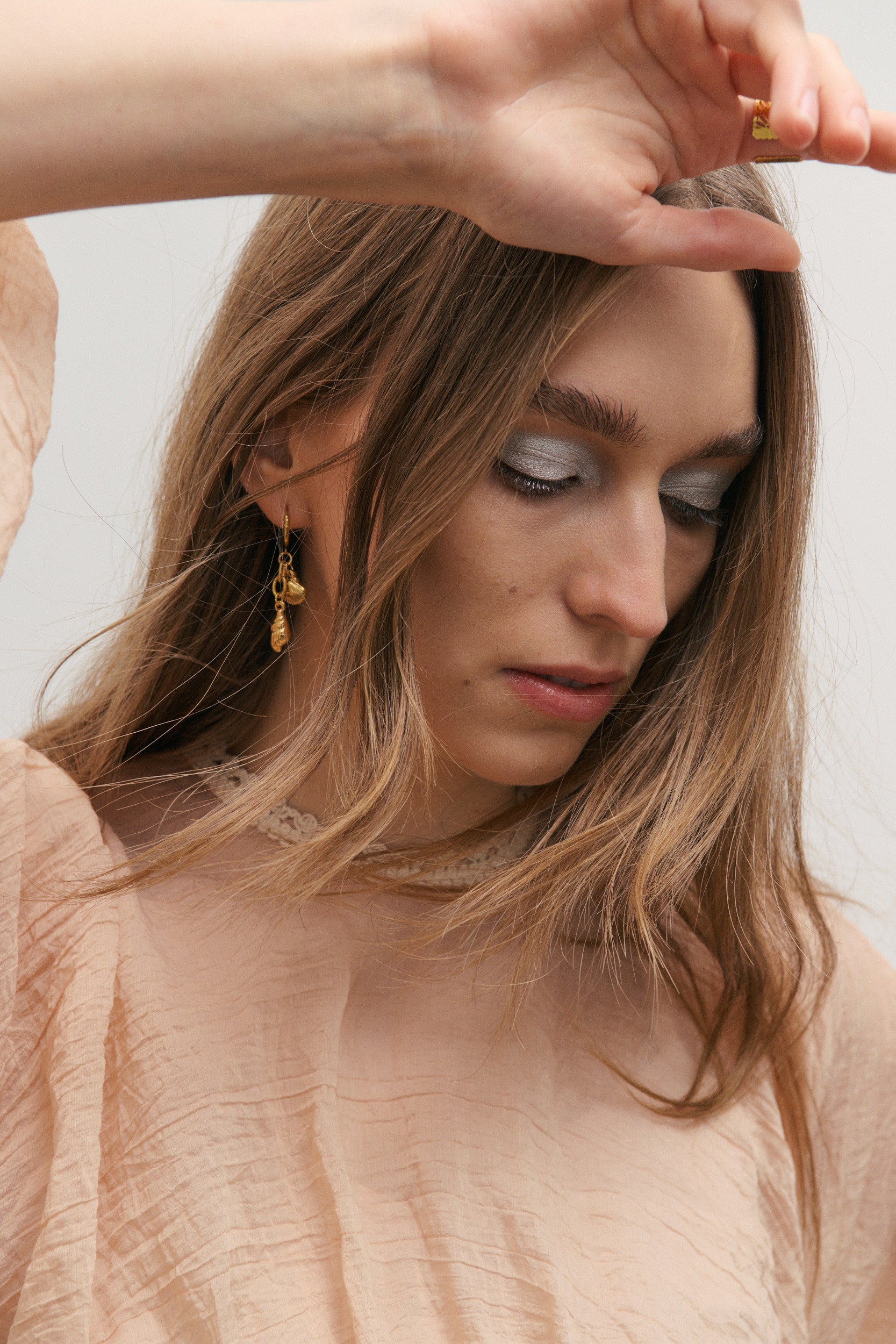 Woman with gold earrings adjusting her hair wearing a light pink top with cream embroidery neck detail against a neutral background