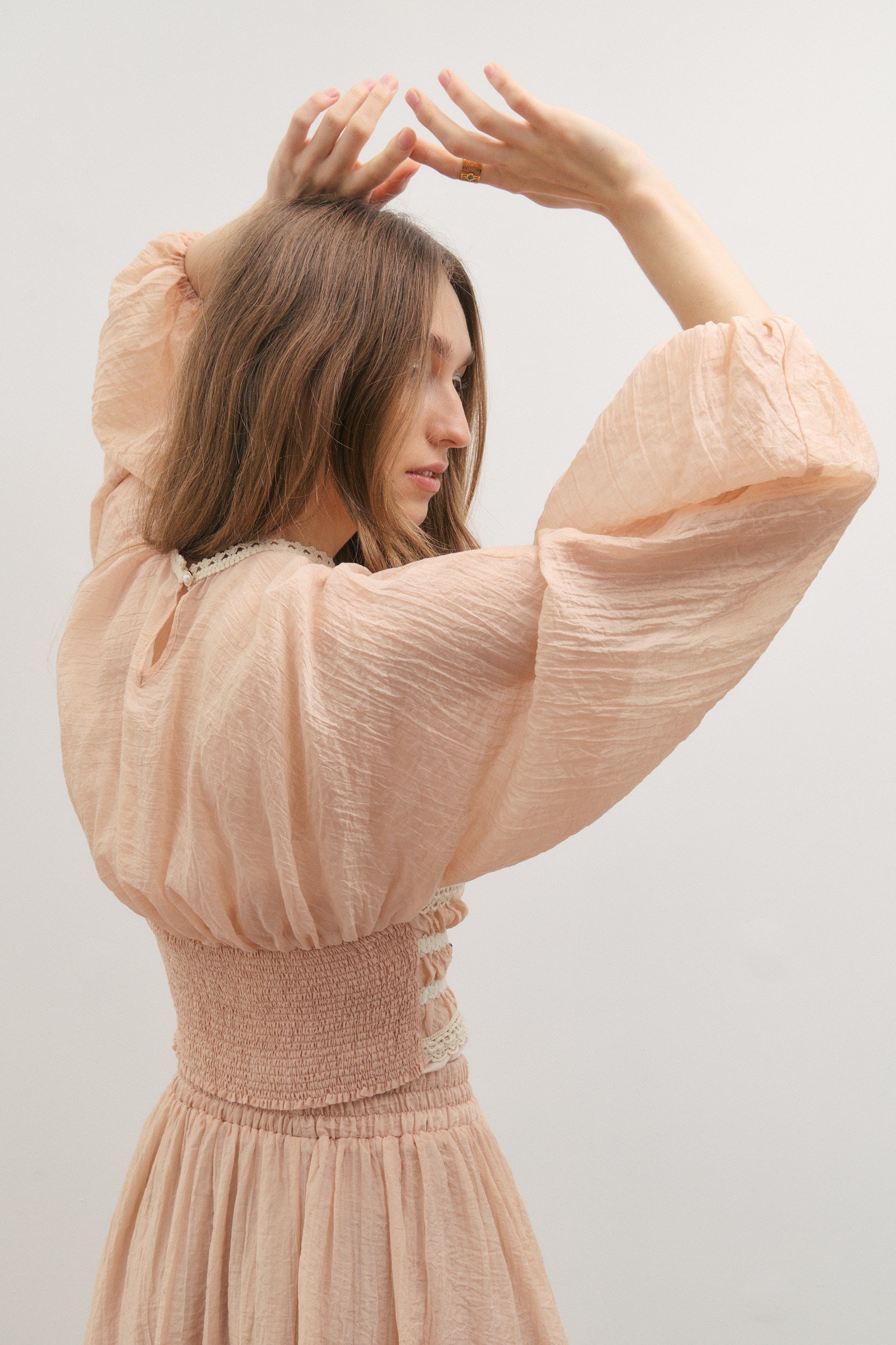 Woman wearing a light pink top with long billowing sleeves with a smocked bodice at the back and cream embroidery detail on the front by fashion brand Maevarii, against a plain background