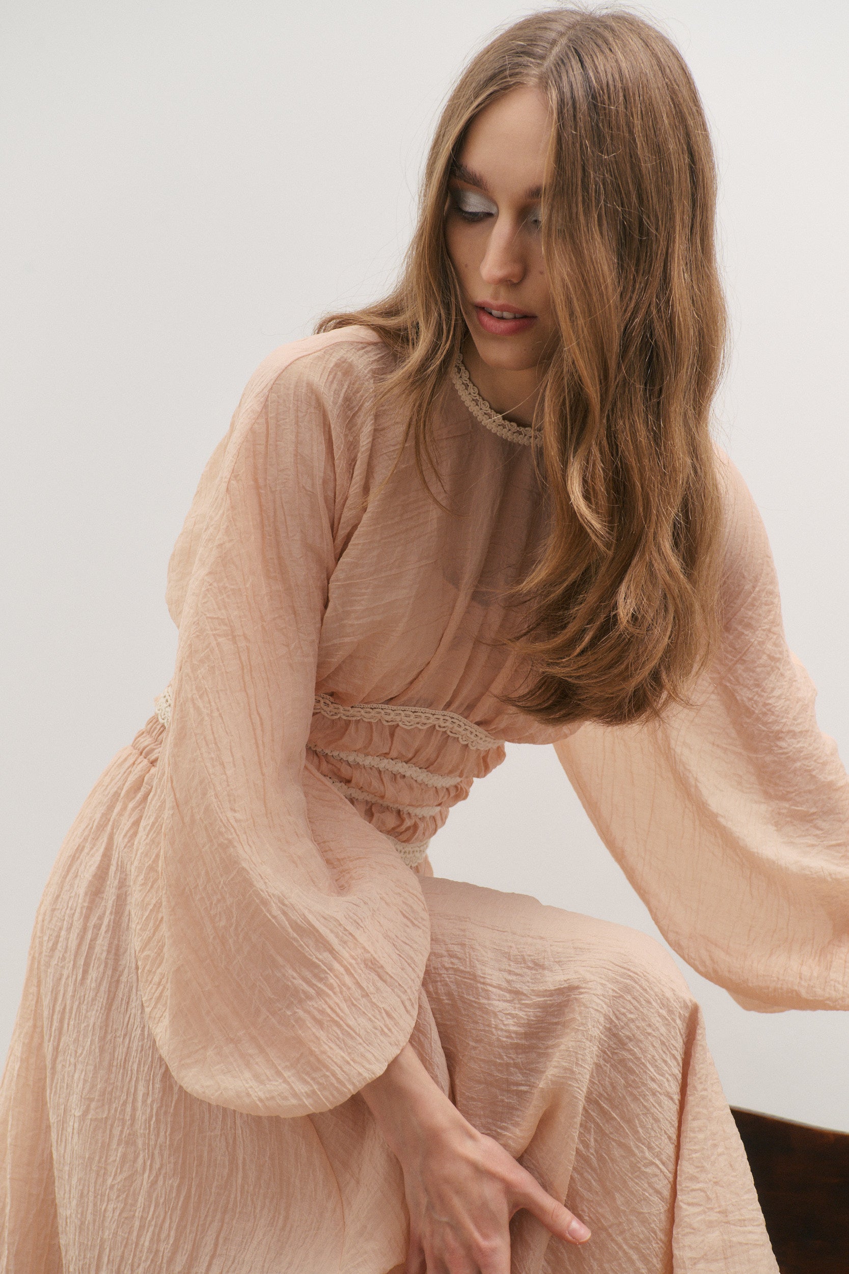 Woman wearing a light pink top named Poetic Dreams for its dreamy whimsical and romantic billowing sleeves and soft colours, sitting against a plain background