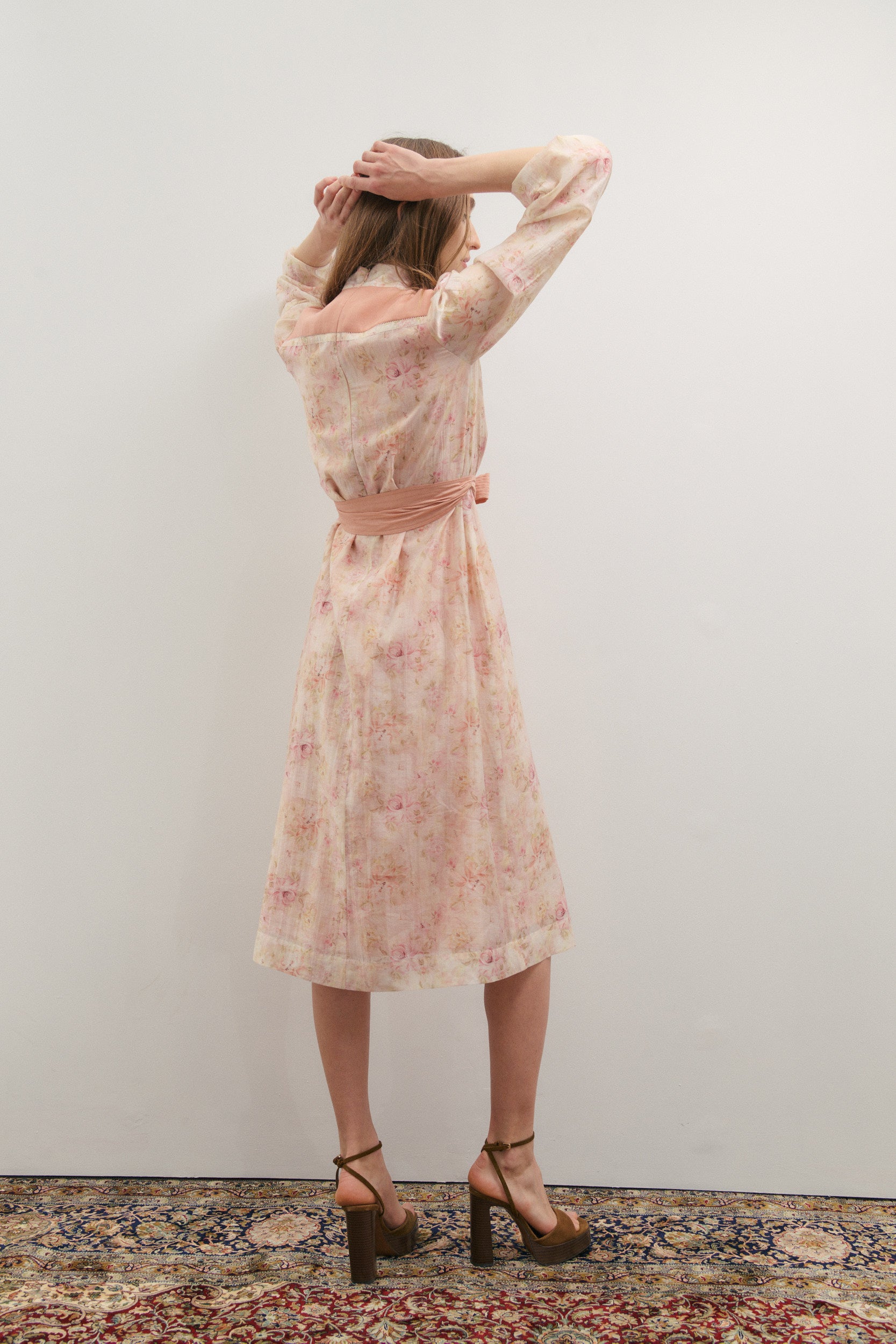 Woman wearing a floral dress standing against a plain wall with a patterned rug on the floor.