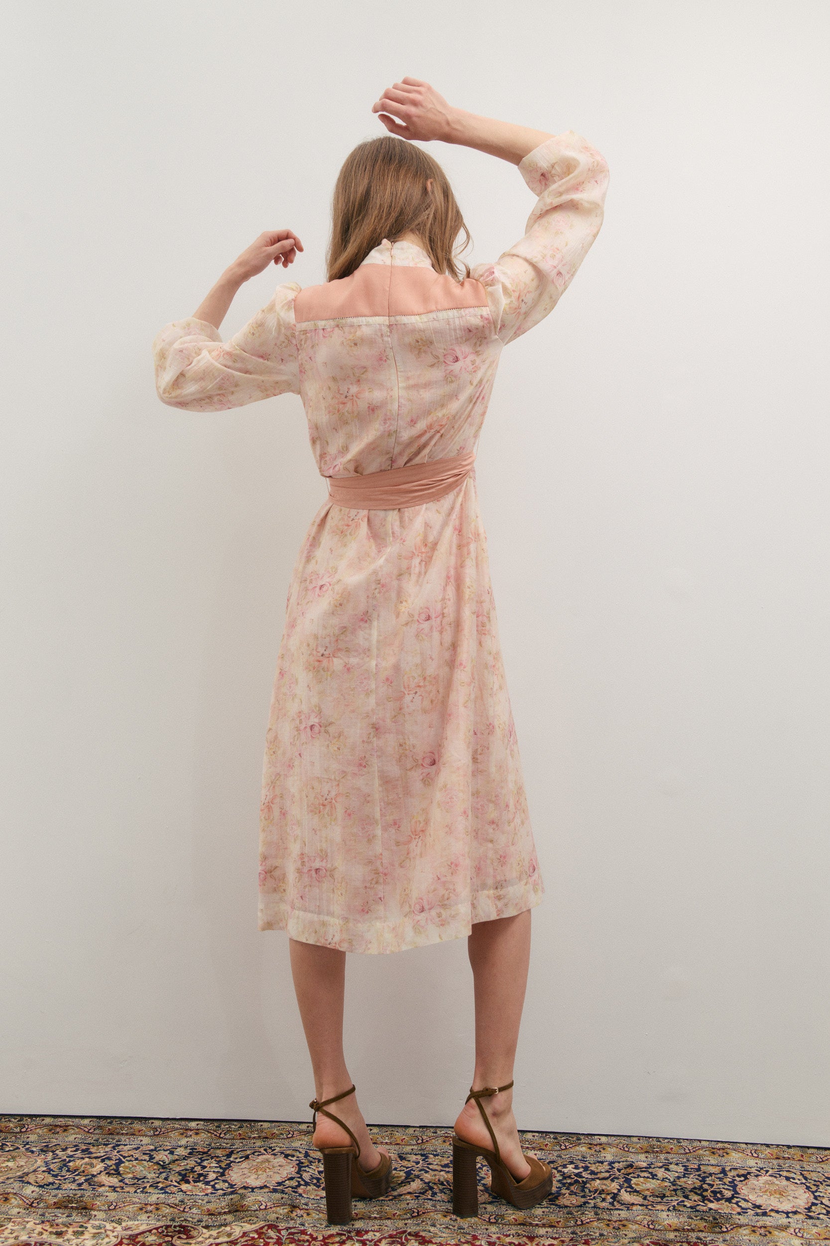 Woman wearing a floral dress with a belt, standing against a plain wall.