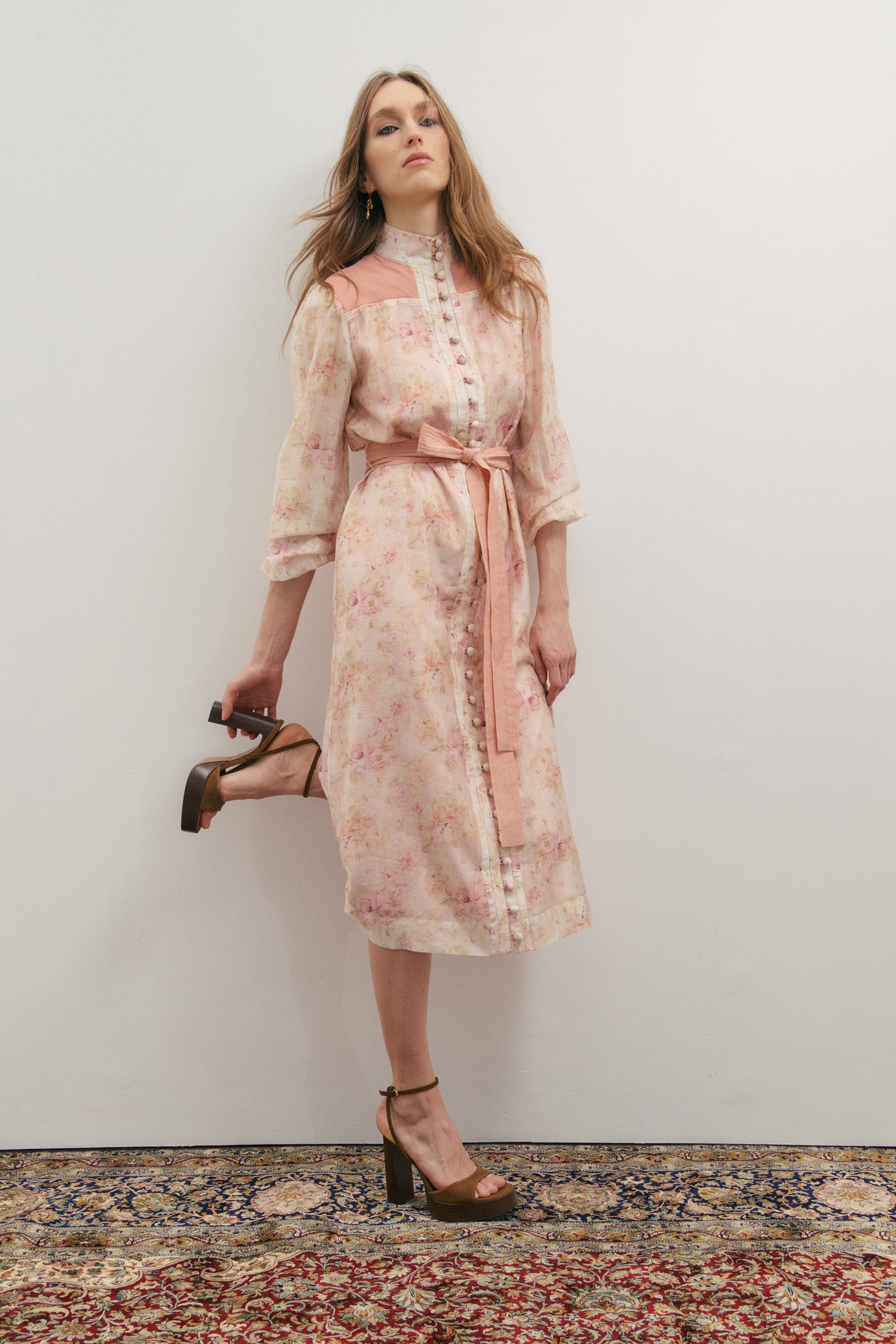 Woman wearing a floral dress with a belt, standing against a plain wall.