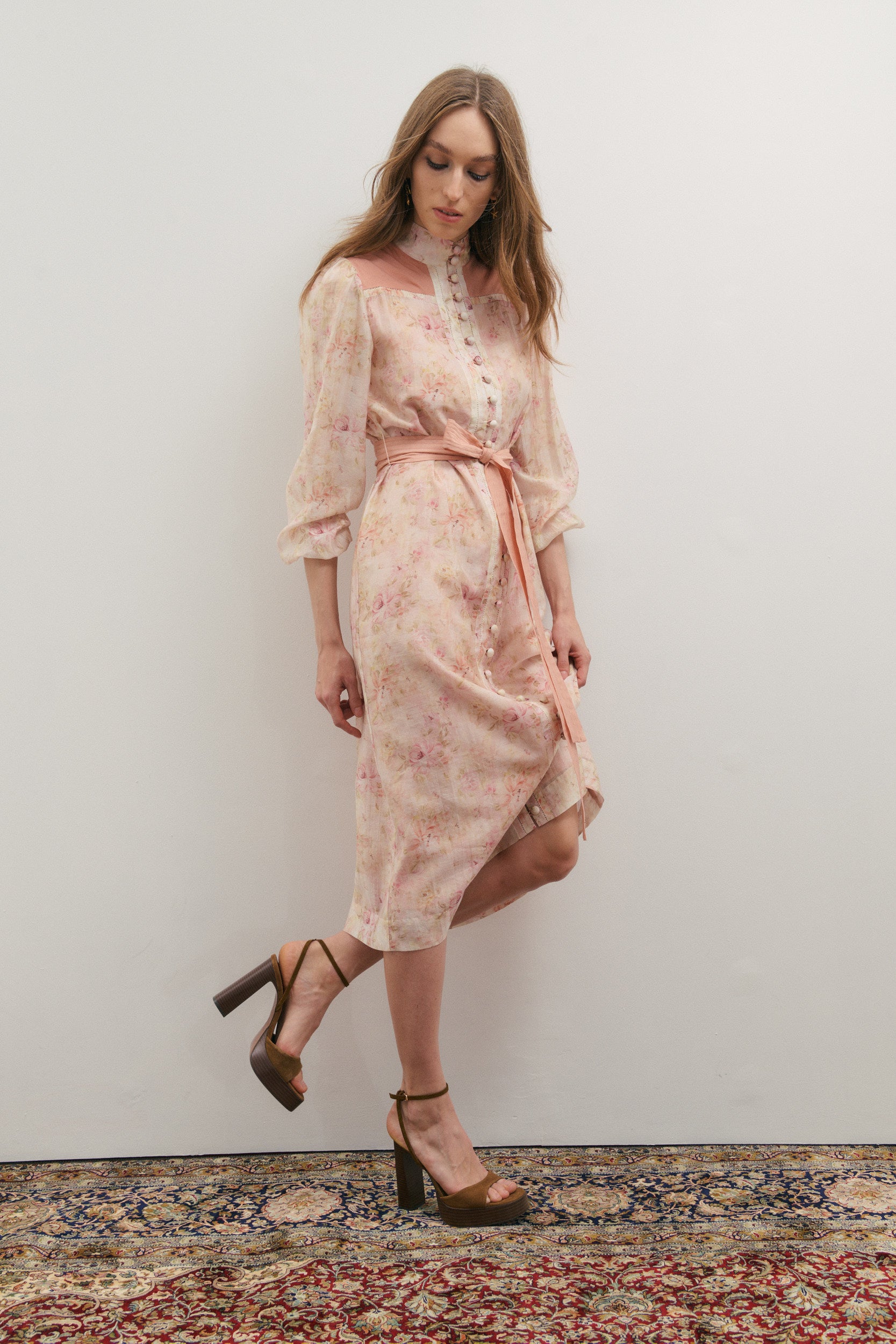 Woman wearing a floral dress standing on a patterned rug against a plain wall.